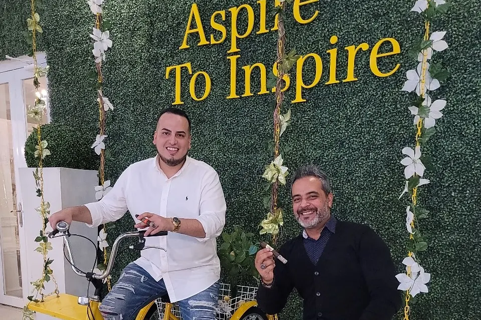 Sergio y Nelson Rivera con cigarros, frente a un muro verde que dice Aspire To Inspire